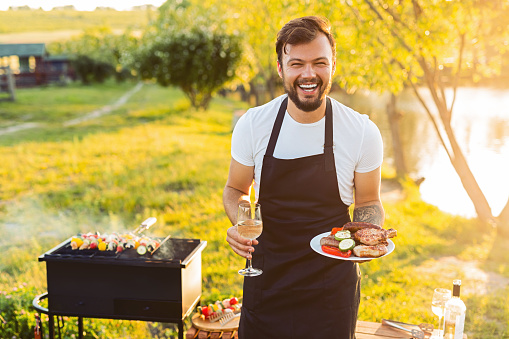 Joyful young bearded guy in t shirt and apron laughing and looking at camera while standing on grassy lawn near lake with plate of grilled meat and veggies and wineglass