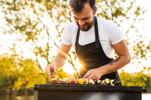 Crop cheerful young ethnic bearded male chef in apron preparing delicious veggie kebabs on grill and smiling during picnic in nature on sunny day