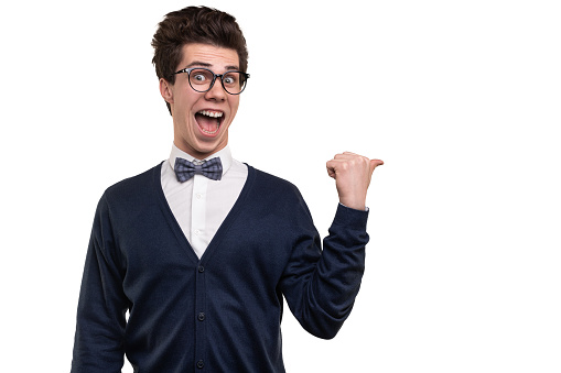 Funny young man in glasses looking at camera with opened mouth and pointing away with thumb during studies against white background