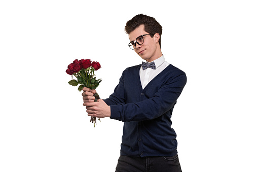 Young intelligent male with dark hair in classy clothes and eyeglasses smiling while standing against white background with bouquet of red roses in hands