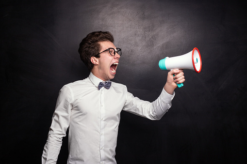 Mad young man in white shirt with bow tie and glasses screaming into megaphone while making announcement against black background