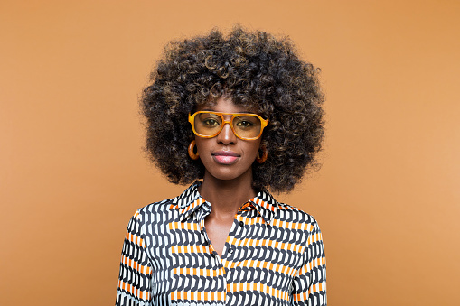 Beautiful young woman with afro hairstyle wearing printed dress, brown glasses and wooden earrings looking at camera. Studio shot on brown background.