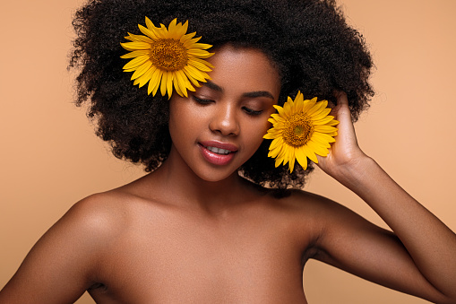Content young African American female millennial with perfect skin and bare shoulders touching dark curly hair with yellow sunflowers and smiling in beige studio