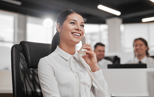 Cheerful young female call center agent with long dark hair in white shirt smiling while talking on telephone during work in modern office with happy male colleagues