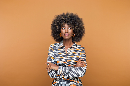 Beautiful young woman with afro hairstyle wearing printed dress and wooden earrings looking away and smiling. Studio shot on brown background.