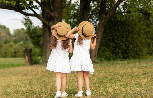 Back view of twin sisters in similar white dresses touching straw hats and admiring tall trees on summer weekend day in park