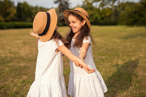 Adorable cheerful little twin girls with long dark hair in similar white dresses and straw hats smiling brightly while playing together on grassy meadow during holidays in countryside
