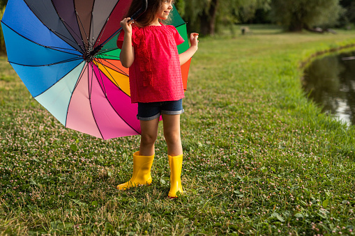 Crop positive little girl with long dark hair in stylish outfit and yellow gumboots smiling and looking away while standing on grassy meadow near pond with colorful umbrella in hand