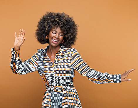 Beautiful young woman with afro hairstyle wearing printed dress and wooden earrings dancing against brown background. Studio shot.