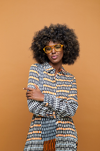 Confident young woman with afro hairstyle wearing printed dress, brown eyeglasses and wooden earrings standing with arms crossed and looking at camera. Studio shot on brown background.