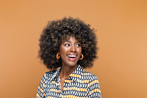 Surprised young woman with afro hairstyle wearing printed dress and wooden earrings looking away and laughing. Studio shot on brown background.