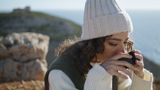 Adventure woman drink coffee on ocean cliff picnic closeup. Serene morning time. Thinking traveler enjoying warm beverage admire view on spring weekend. Pretty curly girl looking camera relax outdoors