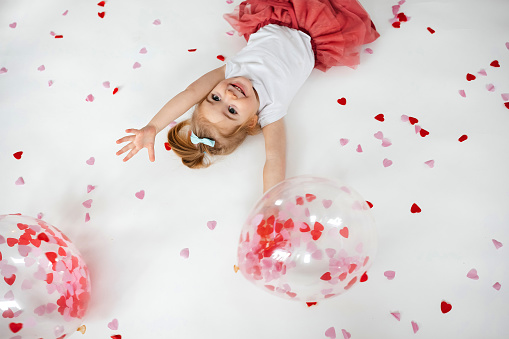 Adorable little girl lying on white background with balloons with heart shape papers inside it and around. Valentines Day concept