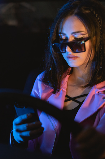 Female portrait of woman who wear sunglasses and using mobile phone indoors the car. Film Noir Style and cyberpunk concept photography, cinematic photo with neon light. People and technology in vehicle.