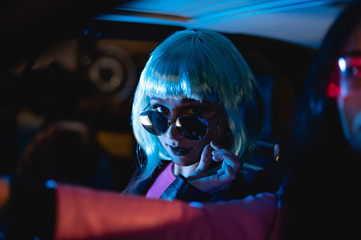Female portrait of two women with blue hairstyle who sitting on the backseat of car. Neon light concept photography, girl is passenger, futuristic photo with color lighting indoors the vehicle