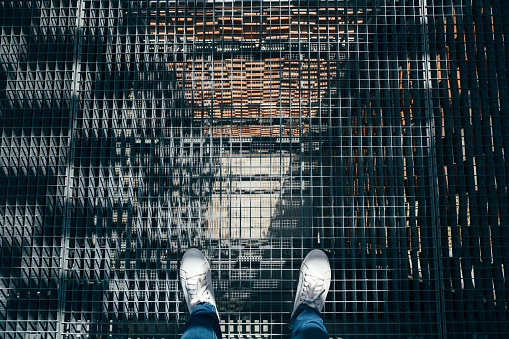 Feet in fashionable white sneakers standing on a metal steel mesh floor in prison, emergency exit staircase, factory, industrial production etc. Man in a steel cage.