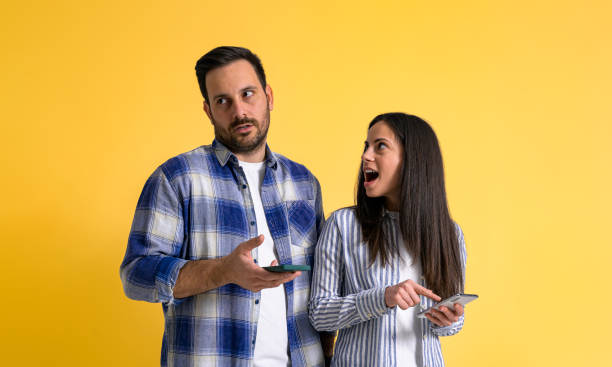 fidanzata sorpresa con la bocca aperta usando il telefono cellulare e guardando il fidanzato maleducato roteando gli occhi irritato e ignorandola su uno sfondo giallo isolato - mouth open women displeased anger foto e immagini stock