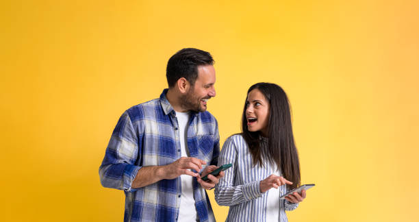 istock Excited and surprised girlfriend with mouth open using smart phone and looking at cheerful boyfriend while standing isolated against yellow background 1475846414