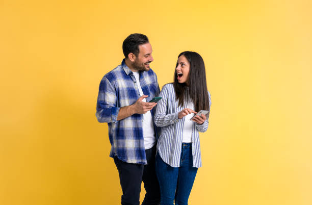 novia feliz y sorprendida con la boca abierta usando el teléfono móvil y mirando al novio alegre mientras está de pie sobre un fondo amarillo aislado - standing digital tablet couple love fotografías e imágenes de stock