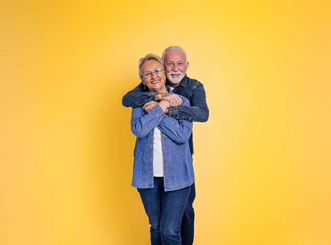 Portrait of romantic loving bearded senior man embracing wife from behind and looking at camera while posing together against yellow background