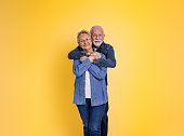 Portrait of romantic loving bearded senior man embracing wife from behind and looking at camera while posing together against yellow background