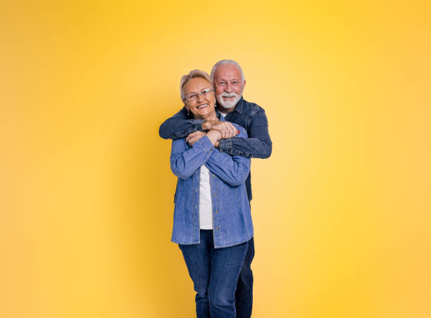 portret romantycznego, kochającego, brodatego starszego mężczyzny obejmującego żonę od tyłu i patrzącego w kamerę, pozując razem na żółtym tle - couple isolated heterosexual couple cheerful zdjęcia i obrazy z banku zdjęć