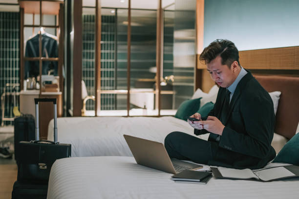 istock Asian businessman replying text message with mobile phone in hotel room sitting on bed with laptop 1475861376