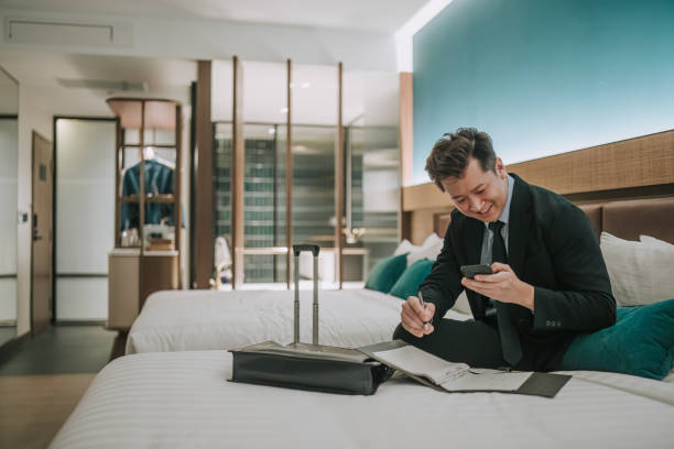 istock Asian businessman working in hotel room sitting on bed writing notes reading phone text messages 1475862074