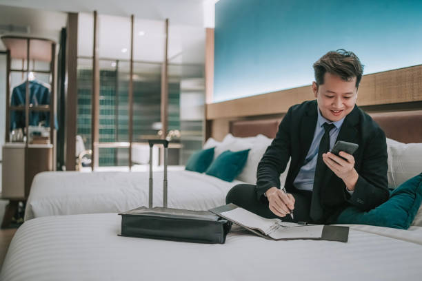 istock Asian businessman working in hotel room sitting on bed writing notes reading phone text messages 1475863134