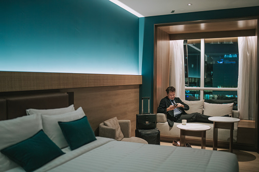 Asian Businessman reading smart phone text message sitting on sofa with luggage and suitcase in hotel room sofa at night