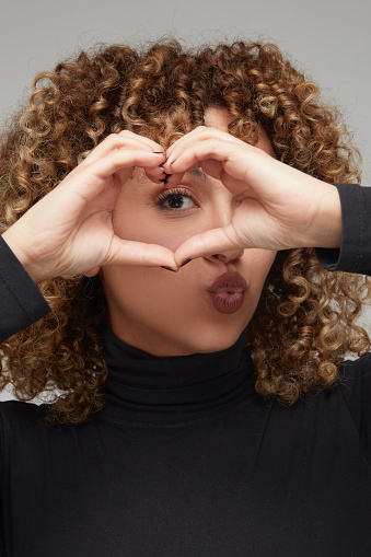 Portrait of beautiful woman with curly hair making heart sign with hands in front of her eyes.