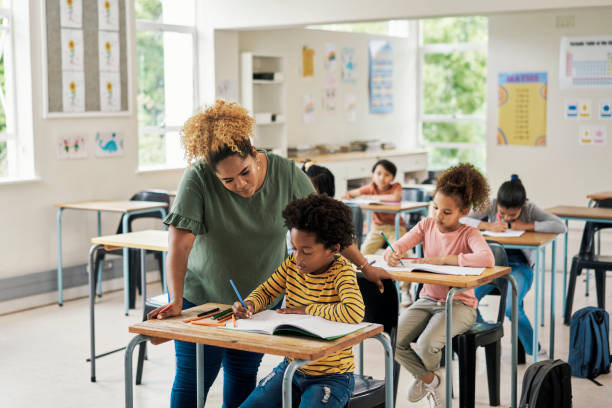 Children classroom help, teacher and test writing of kids in a exam lecture hall for knowledge. Education explanation, student learning and school employee in a study lesson with a young boy working stock photo