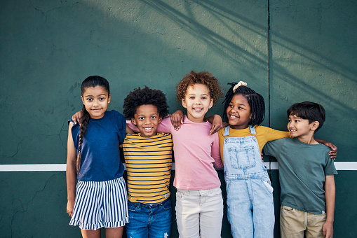 Diversity, friendship and portrait of children on a playground hugging after playing together. Happy, smile and multiracial young kid friends standing, embracing and bonding by a wall on park in city