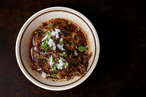 A bowl of slow cooked and braised birria meat in a vintage bowl on a dark brown background with copy space.