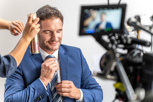 Television studio assistant preparing businessman for broadcast and using hairspray