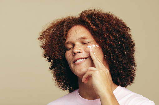 Young man with curly ginger hair gets into his skincare routine, applying beauty cream to his freckled face. Man with unique beauty features pampers his skin to maintain his healthy and youthful glow.