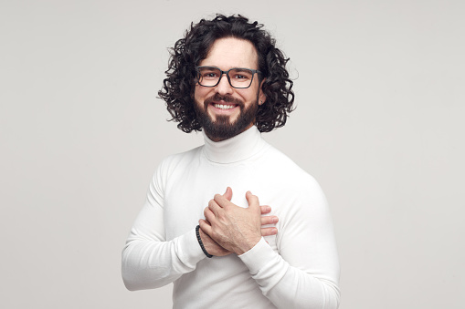 Happy adult male model in eyeglasses holding hands on chest feeling grateful and thankful while looking at camera with gentle smiling face