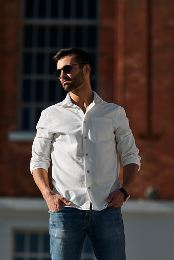 Confident young stylish ethnic male with dark hair and beard in trendy sunglasses and smart casual outfit standing on street with hands in pockets and looking away on sunny day