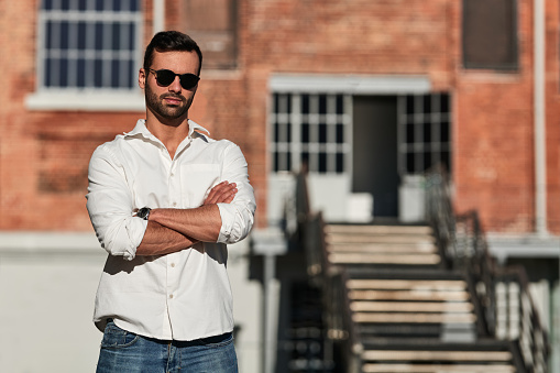 Self assured young Arab male entrepreneur with dark hair and beard in white shirt and sunglasses standing on street against brick building with folded arms on sunny day