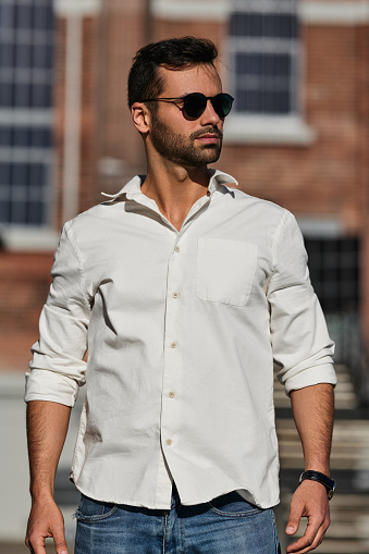 Self assured male in white shirt and sunglasses looking away while standing on blurred background of city street in daytime