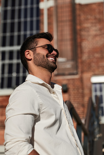 Happy ethnic male in white shirt and stylish sunglasses leaning on railing and smiling while resting outside brick building on sunny day