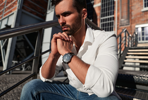 Low angle of unhappy ethnic male in white shirt with wristwatch clasping hands and pondering while sitting on steps on sunlit city street