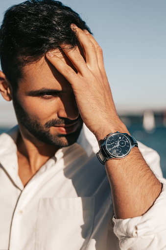 Handsome ethnic male in white shirt with wristwatch touching dark hair and looking away on blurred background of river and blue sky