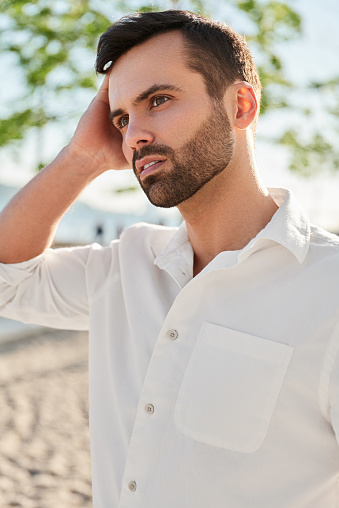 Handsome young male tourist in white shirt touching dark hair and looking away while resting on blurred background of beach on sunny summer day on resort