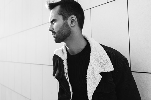 Black and white pensive bearded male in jacket with fur collar frowning and looking away while leaning on tiled wall on city street