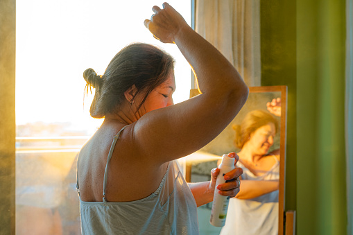 Portrait of  mature woman applying deodorant on underarm in front of a mirror during morning time. High resolution 42Mp indoors digital capture taken with SONY A7rII and Zeiss Batis 40mm F2.0 CF lens