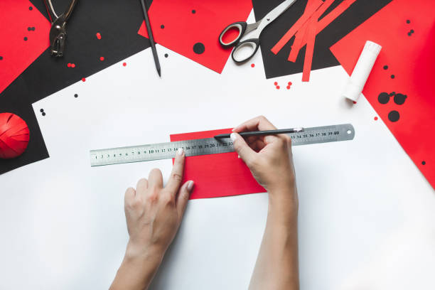 How to make paper red and black ladybug toy with children at home. Step by step instructions. Hands making DIY summer project. Top view. Step 3. On sheet of red paper 15 cm long, draw 10 lines 1 cm wide