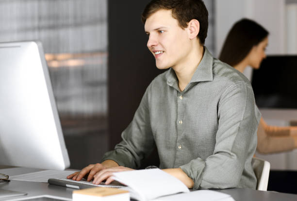 istock Dark-haired young businessman and programmer in a green shirt is working hard on his computer, while sitting at the desk in a modern cabinet with a female colleague on the background. Concept of successful business start up 1475927086