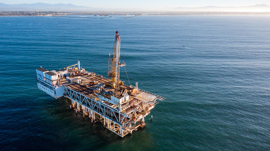 Off the coast of Long Beach, California, an imposing offshore drilling rig rises from the waves, a towering structure of steel and machinery. The rig is a hive of activity, with workers toiling around the clock to extract oil and gas from beneath the ocean floor. The sound of heavy machinery fills the air, and the smell of petroleum lingers on the sea breeze.\n\nThe rig is a marvel of engineering, with massive cranes and drills capable of reaching depths of thousands of feet below the surface. It is a symbol of America's reliance on fossil fuels, and the risks and rewards that come with offshore drilling.\n\nDespite its imposing size, the rig is a small speck in the vastness of the ocean, and its impact on the surrounding ecosystem is a matter of ongoing debate. Proponents argue that the benefits of domestic energy production outweigh the potential environmental risks, while opponents point to the dangers of oil spills and the long-term impact on marine life.\n\nAs the sun sets on the horizon, the rig continues to churn away, a reminder of the complex and often contentious relationship between humans and the natural world.