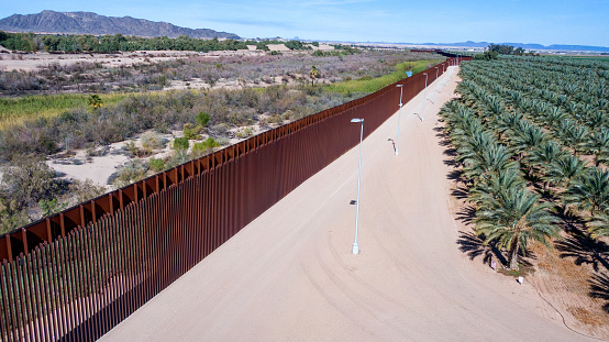 The drone picture captures the stark contrast between the imposing American border wall and the tranquil field of palm trees in Yuma, Arizona. The wall, with its tall steel bars, dominates the foreground of the image, a symbol of the ongoing debate over immigration policy in the United States. In contrast, the palm trees offer a serene and calming sight, reminding us of the natural beauty that exists alongside human-made structures. The image also highlights the contentious nature of the border wall and its impact on local communities and ecosystems. Despite the stark differences between the wall and the palm trees, both are part of the complex and evolving landscape of the US-Mexico border region.